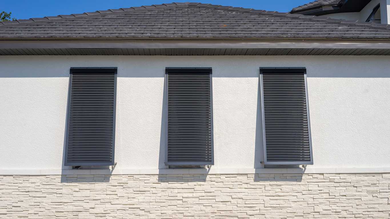 Three storm shutters, hinged at the top, over three tall windows on a house in Florida
