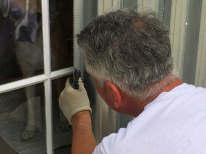Man installing shutters over window while dog watches from inside house